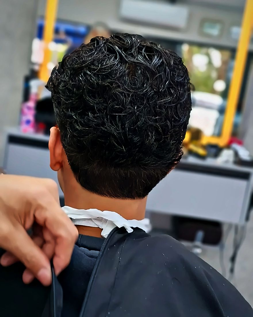 A nuca de um cliente, cabelo cacheado, com a mão do barbeiro ajeitando a capa